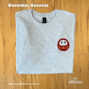May include: A light gray t-shirt with a Daruma doll design on the front. The Daruma doll is red with gold accents and the Japanese character for "fortune". The text "Daruma: Success" is at the top. The brand name "Crafty Kazoku" is at the bottom.