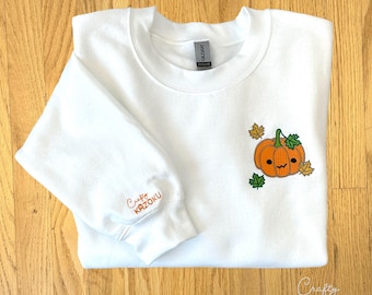 以下が含まれることがあります： オレンジ色のカボチャのデザイン、緑の葉、そして「Pumpkin」の文字が刺繍された白いスウェットシャツ。「Crafty Kazoku」の文字が袖にあります。画像の下部には「Crafty Kazoku」の文字があります。
