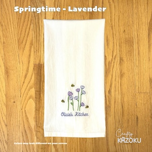 Pode incluir: Toalha de cozinha branca com bordados de flores de lavanda e abelhas. O texto "Olivia's Kitchen" é bordado abaixo. O texto "Springtime - Lavender" está no topo.
