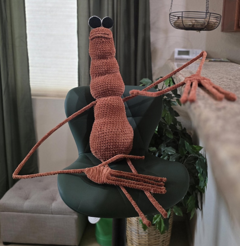 Puede incluir: Una mu&ntilde;eca de insecto de ganchillo marr&oacute;n hecha a mano con grandes ojos negros y largas y delgadas patas. El peluche est&aacute; sentado en una silla verde oscuro, con los brazos extendidos. El cuerpo del insecto est&aacute; segmentado y la mu&ntilde;eca tiene una apariencia caprichosa y de dibujos animados.