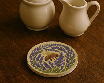 May include: A round coaster with a bee and lavender design. The coaster features a tan background with a border of purple lavender and green leaves. A brown and yellow bee is in the center. A sugar bowl and creamer are in the background.