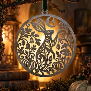 May include: A circular metal ornament featuring a laser-cut design of a rabbit standing among foliage, mushrooms, and pumpkins. Above the rabbit is a crescent moon with a star. The ornament hangs from a dark green velvet ribbon loop.