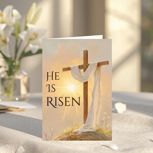 May include: A folded greeting card with the words "He Is Risen" printed on the front. The card features a painting of a wooden cross draped with a white cloth, set against a sunrise. A vase of white lilies sits in the background.