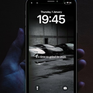 Può includere: Uno smartphone con uno sfondo in bianco e nero di auto sportive di lusso. Lo schermo mostra la data, l'ora e la frase "It's time to grind its 2026". Il telefono è tenuto in mano su uno sfondo scuro.