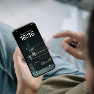 May include: A black smartphone displays a dark wallpaper featuring a black vehicle. The screen shows the time 18:36 and the date Wednesday, 13 August. The phone is held in someone's hands.