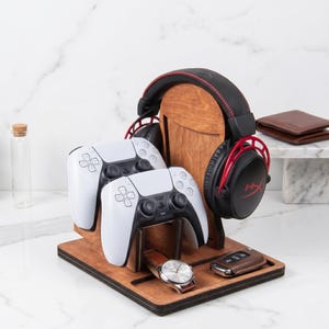 Puede incluir: Un soporte organizador de madera para accesorios de juego que incluye un soporte vertical para auriculares negros y rojos, dos mandos de juego inal&aacute;mbricos blancos, un reloj anal&oacute;gico plateado con correa de cuero marr&oacute;n y un llavero de cuero marr&oacute;n.