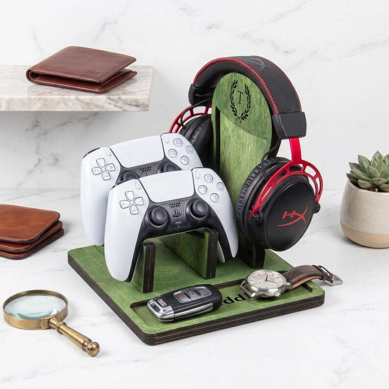 Puede incluir: Un organizador de juegos de madera verde con un soporte vertical grabado con una corona de laurel y la letra T. Sostiene dos mandos de juego blancos y unos auriculares negros y rojos. La base incluye una bandeja para objetos peque&ntilde;os.