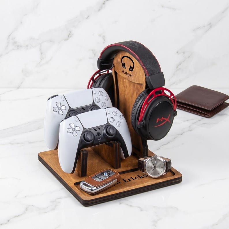 Puede incluir: Un soporte organizador de madera para juegos con un soporte vertical para auriculares con el texto Robyn, dos mandos de juego blancos, un llavero de coche de cuero marr&oacute;n, un reloj anal&oacute;gico plateado y el nombre Ericksen grabado en la base.