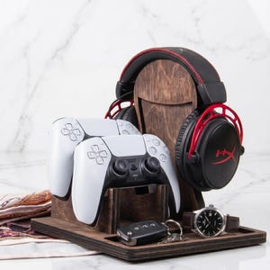 Puede incluir: Un soporte organizador de madera oscura para accesorios de juego, con una base vertical para auriculares negros y rojos, un soporte frontal para dos mandos blancos y una base plana con una llave de coche negra y un reloj de pulsera con correa marr&oacute;n.