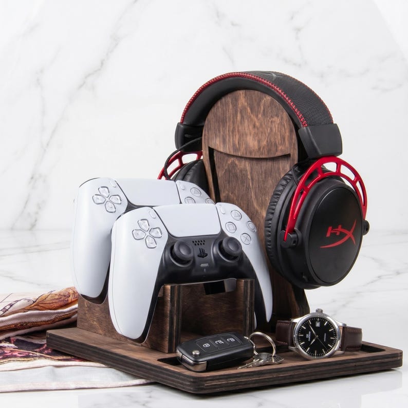 Puede incluir: Un soporte organizador de madera oscura para accesorios de juego, con una base vertical para auriculares negros y rojos, un soporte frontal para dos mandos blancos y una base plana con una llave de coche negra y un reloj de pulsera con correa marr&oacute;n.