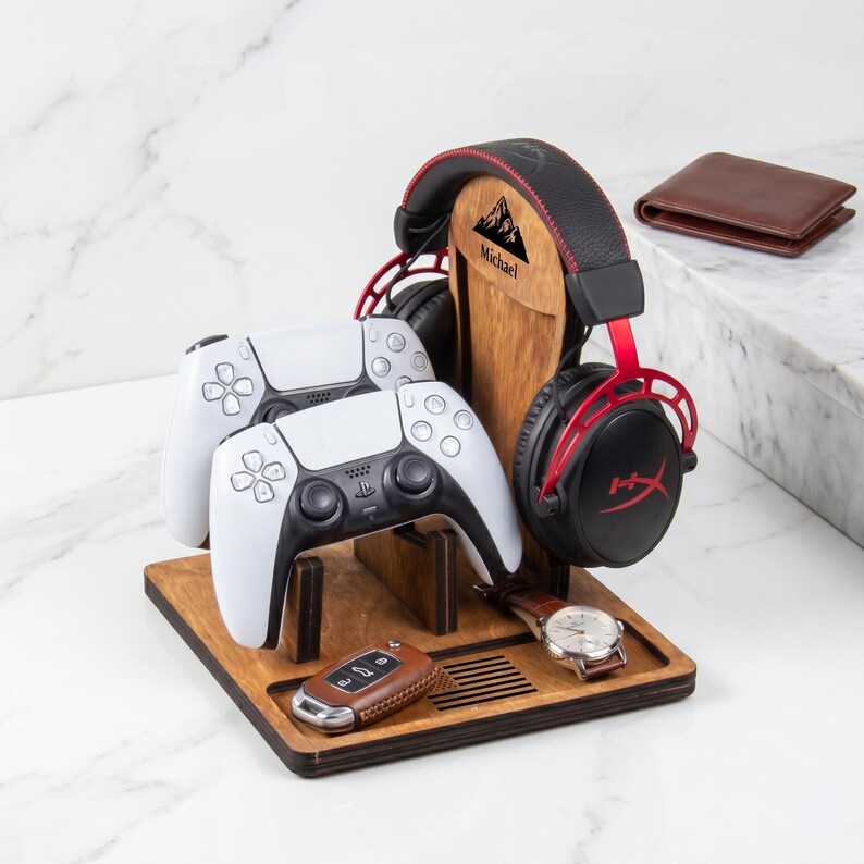 Puede incluir: Un soporte organizador de madera para juegos con un montaje vertical para auriculares, dos ranuras para mandos y una bandeja base. El soporte est&aacute; grabado con un gr&aacute;fico de monta&ntilde;a y el nombre Michael. Contiene auriculares negros y rojos, dos mandos blancos, un llavero de cuero marr&oacute;n y un reloj de pulsera plateado.