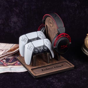 Pode incluir: Uma estação de jogos de madeira com dois controles brancos e pretos, um fone de ouvido preto e vermelho e o nome "Robert Lloyd" gravado. O fone de ouvido repousa em um suporte de madeira com um design decorativo.