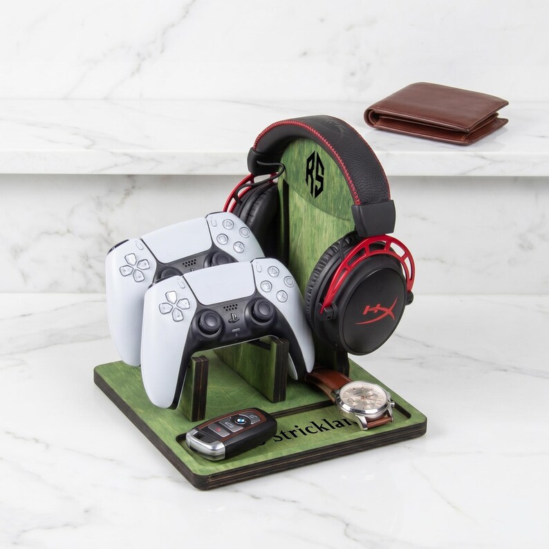 Puede incluir: Un soporte organizador de juegos de madera verde con un soporte vertical con un logotipo RS, que sostiene dos mandos blancos, unos auriculares negros y rojos, un reloj de pulsera y una llave de coche, con el nombre Strickland grabado en la base.