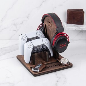 May include: A dark wood gaming organizer stand featuring a vertical headphone mount with an engraved wreath and initials R M, two white game controllers, a brown leather key fob, a silver wristwatch, and the engraved name Robert Lloyd.