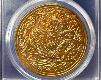 Könnte beinhalten: Eine Goldmünze in einem transparenten Kunststoffgehäuse. Die Münze zeigt ein detailliertes Drachendesign. Der Halter hat ein Etikett mit chinesischen Schriftzeichen, dem Text "MS 65" und den Maßen 39,5*3,0mm und einem Gewicht von 37,5g.