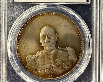 Könnte beinhalten: Eine Silbermünze in einem transparenten Kunststoffgehäuse. Die Münze zeigt ein Porträt eines Mannes in Uniform und den Text "CHINA-REPUBLIC". Das Gehäuse hat ein Etikett mit dem Text "NCAC AU 55" und "NUMISMATIC CULTURAL INNOVATION".