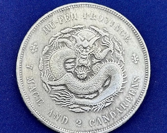May include: A silver coin featuring a detailed dragon design at the center, surrounded by the text "HU-PEH PROVINCE" and "7 MACE AND 2 CANDAREENS". The coin has a textured edge and is set against a dark blue background.