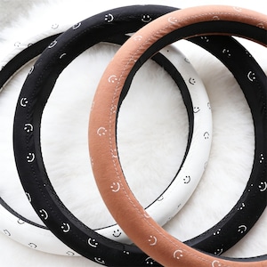 Pode incluir: Três capas de volante em preto, branco e castanho. Cada capa é decorada com pequenas carinhas sorridentes brancas. As capas são feitas de um material macio e são projetadas para caber em volantes padrão.