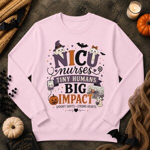 Può includere: Una felpa a maniche lunghe rosa chiaro con una stampa grafica che riporta il testo NICU nurses TINY HUMANS BIG IMPACT SPOOKY SHIFTS STRONG HEARTS. Il design include illustrazioni di fantasmi, una zucca, uno stetoscopio e un'incubatrice.