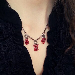 May include: A delicate necklace with a dark metal chain and three clusters of small, red beads. The necklace is worn over a black top with a ruffled neckline and clear buttons. The text "Rock Spirit Studio" is visible.