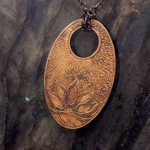 May include: A gold oval pendant necklace featuring a lotus flower design. The pendant has a textured surface and a hole at the top for the chain.