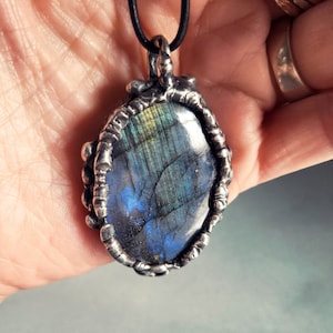 May include: A silver pendant with a blue and green labradorite gemstone set in a wire-wrapped bezel. The pendant is hanging from a black cord.