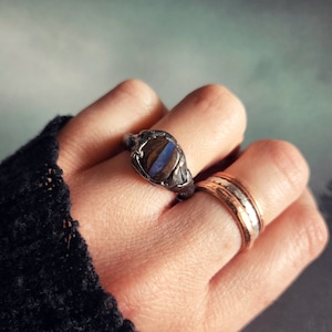May include: A silver ring with a blue and brown opal stone set in a textured band. The ring is worn on a hand with other rings.