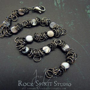 May include: A handcrafted bracelet featuring a chainmail design with dark gray metal rings and iridescent, round, and flat beads. The bracelet has a lobster clasp closure. The Rock Spirit Studio logo is visible in the bottom right corner.