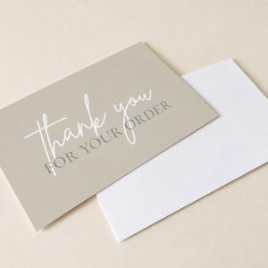 Può includere: Un cartoncino rettangolare color beige con la scritta in corsivo bianco thank you e il testo grigio con grazie FOR YOUR ORDER, che si sovrappone parzialmente a una busta rettangolare bianca. Il cartoncino e la busta sono posizionati in diagonale.