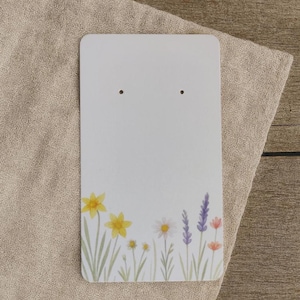 Könnte beinhalten: Eine weiße Ohrring-Präsentationskarte mit zwei Löchern oben. Der untere Teil der Karte zeigt ein Aquarell-Blumenmuster mit gelben Narzissen, weißen Gänseblümchen, lila Lavendel und rosa Blüten. Die Karte liegt auf einem beigen Leinentuch.