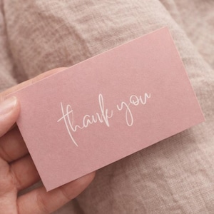 May include: A rectangular dusty pink card featuring the words thank you printed in a white cursive script font. The card has a smooth matte finish and is held against a textured fabric background of a similar muted pink colour tone.