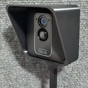 May include: A dark gray Blink security camera mounted inside a protective housing with an integrated sun visor. The camera unit features a central lens and sensor, with the brand name Blink printed in white text at the bottom of the front panel.