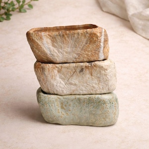 Könnte beinhalten: Drei rechteckige Pflanzgefäße, die übereinander gestapelt sind. Das obere Pflanzgefäß ist hellbraun mit weißen Streifen, das mittlere ist hellbeige und das untere ist hellgrün mit einem gesprenkelten Muster. Die Pflanzgefäße haben ein raues, strukturiertes Aussehen. Jedes Pflanzgefäß ist etwa 20 cm lang.