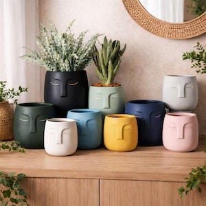 以下が含まれることがあります： 黒、緑、青、黄色、白、ピンクなど、さまざまな色の顔型セラミックプランターのコレクション。各プランターは、閉じられた目と鼻を持つミニマリストの顔のデザインが特徴です。一部のプランターには植物が入っています。