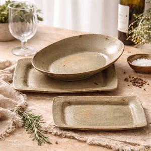 May include: A collection of handcrafted ceramic tableware in green and brown tones. The set comprises an oval bowl, two rectangular plates, and a small bowl. The plates and bowl have a textured finish, giving them a natural, earthy appearance.