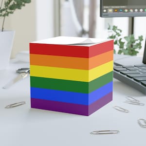 May include: A cube-shaped notepad with a rainbow design, featuring horizontal stripes of red, orange, yellow, green, blue, and purple. The notepad sits on a white surface with scattered silver paper clips.