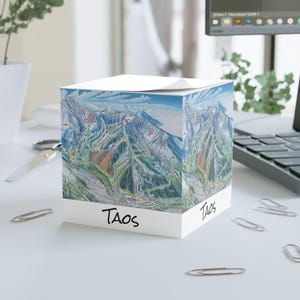 May include: A white cube-shaped notepad featuring a detailed map of a mountain ski resort. The map is in shades of blue, green, and brown. The word "TAOS" is printed in black on the front of the notepad.