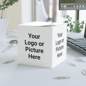 Puede incluir: Una caja blanca en forma de cubo con el texto "Your Logo or Picture Here" impreso en negro en dos lados. Varios clips plateados están esparcidos alrededor de la caja sobre una superficie blanca. Un par de tijeras y un portátil son visibles en el fondo.