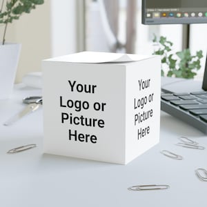 Puede incluir: Una caja blanca en forma de cubo con el texto "Your Logo or Picture Here" impreso en negro en dos lados. Varios clips plateados están esparcidos alrededor de la caja sobre una superficie blanca. Un par de tijeras y un portátil son visibles en el fondo.
