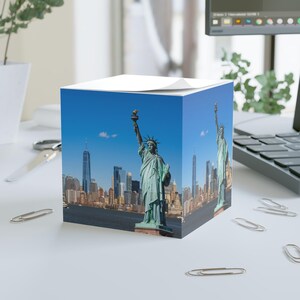 Pode incluir: Um bloco de notas quadrado com uma fotografia da Estátua da Liberdade e o horizonte da cidade de Nova York sob um céu azul claro. O bloco de notas é branco no topo e está sobre uma superfície branca com clipes de papel.