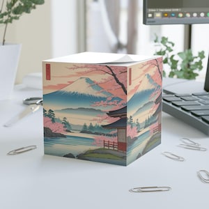 以下が含まれることがあります： 富士山、桜、伝統的な建物が描かれた日本の風景デザインの正方形のメモ帳。色使いは青、ピンク、ベージュ。メモ帳は、クリップの付いた白い表面に置かれています。