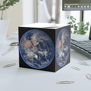 Può includere: Un blocco note a forma di cubo nero con un'immagine dettagliata della Terra su ogni lato. L'immagine della Terra mostra continenti, oceani e nuvole. Il blocco note è su una superficie bianca con graffette sparse.