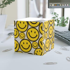 May include: A cube-shaped notepad featuring a repeating pattern of yellow smiley faces with black outlines. The notepad is on a white desk with paper clips, a laptop, and scissors in the background. The design is cheerful.