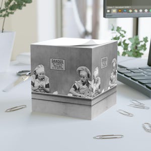 May include: A square paper cube featuring a black and white scene of people in chef uniforms working at a conveyor belt. A sign on the wall reads DANGER BE CAREFUL WHEN BELT IS IN MOTION. The cube has a stack of white paper sheets on the top surface.