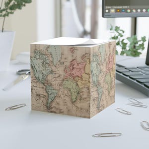 May include: A cube-shaped notepad featuring a vintage world map design. The map is printed in muted colors of green, pink, and beige, with a white top for writing notes. Several silver paper clips are scattered around the notepad on a white surface.