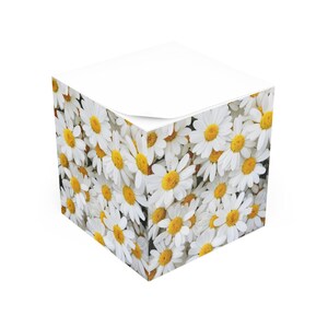 May include: A white cube-shaped notepad covered in a pattern of white daisies with yellow centers. The top of the notepad is white, with a corner of a sheet lifted up. The daisies are densely packed, creating a floral design.