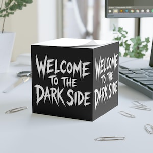 May include: Black sticky note cube with the text "WELCOME TO THE DARK SIDE" in white, stylized lettering. The cube is sitting on a white surface with paper clips scattered around.