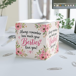 May include: A square sticky note cube featuring a white background with pink floral illustrations and small gold hearts. The text reads, Always remember how much your Besties love you! in black and pink script and sans-serif fonts on the front and side.