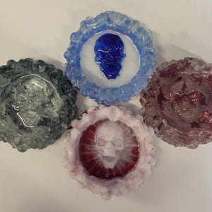 May include: Four decorative ashtrays with skull designs. The ashtrays are in various colors, including clear, blue, red, and white. Each ashtray has a unique design, with the skulls in the center of the ashtrays.