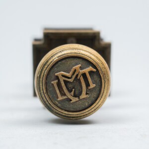 Könnte beinhalten: Ein runder Türknauf aus antikem Messing mit einem erhabenen, geprägten Monogramm mit den Buchstaben IMT in der Mitte. Der Knauf hat einen strukturierten, geriffelten Rand um die kreisförmige Vorderseite und ist an einer quadratischen Platte befestigt.